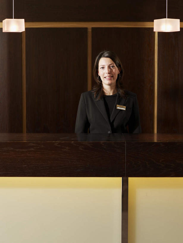 西方人,前台,服务员,站,笑,微笑,竖图,室内,白天,正面,工作服,制服,桌面,桌子,酒店,指示牌,欧洲,欧洲人,木制,标志,文字,墙,注视,标识,吊牌,墙壁,墙面,木制品,胸牌,观察,看,站着,观看,察看,徽章,关注,身份,胸章,胸卡,编号牌,雕刻牌,工号牌,胸号牌,胸徽,员工牌,工作制服,接待员,白种人,服务生,侍者,站立,半身,彩图,工作套服,旅店