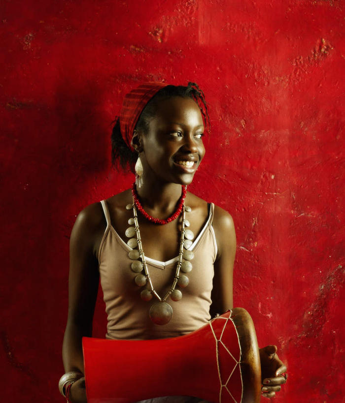 西方人,一个人,站,笑,微笑,露齿笑,竖图,室内,白天,正面,项链,吊带,欧洲,非洲,黑人,欧洲人,仅一个年轻女性,仅一个女性,仅一个人,握,服装,风俗,民俗,墙,鼓,乐器,拿,拿着,扭头,注视,吊带衫,墙壁,墙面,装饰品,衣服,红色,饰品,首饰,观察,看,站着,转头,握着,服饰,观看,打击乐器,察看,节奏,节拍,衣着,穿着,关注,非洲鼓,衣饰,民风,传统乐器,年轻女性,女人,女性,非洲人,站立,半身,彩图