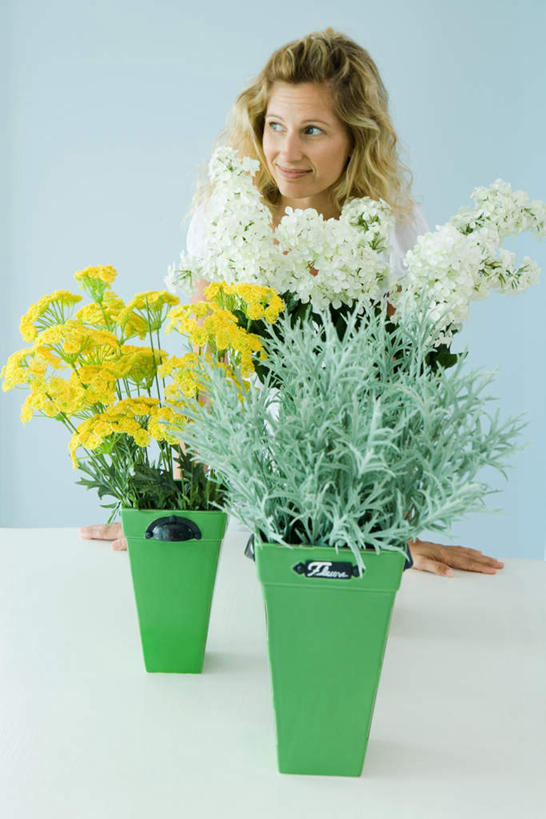 西方人,一个人,站,笑,微笑,露齿笑,竖图,室内,白天,正面,植物,桌面,桌子,叶子,欧洲,欧洲人,仅一个年轻女性,仅一个女性,仅一个人,盛开,绿叶,两束,花,花瓣,花朵,花卉,鲜花,黄色,白色,瓶子,塑料制品,花蕊,站着,嫩叶,塑料瓶,年轻女性,女人,女性,白种人,叶,叶片,站立,半身,彩图