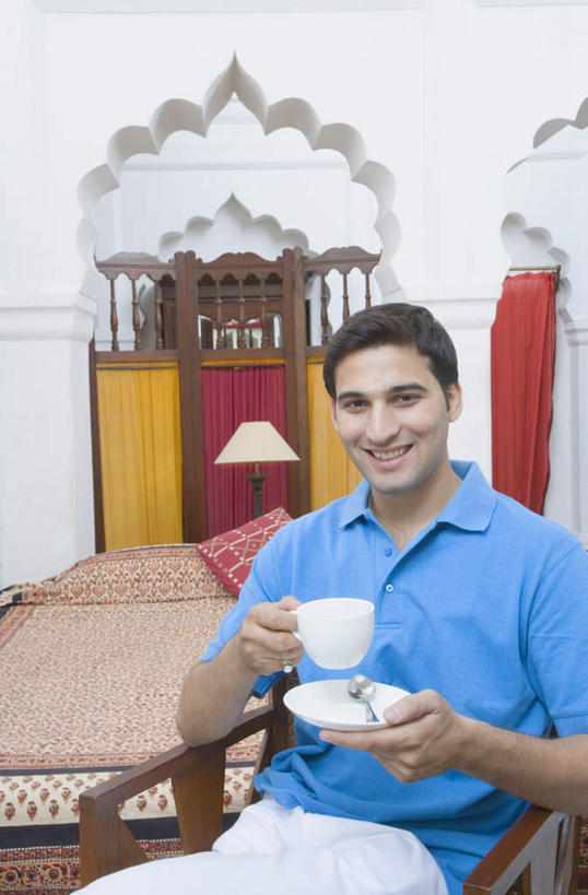 一个人,坐,笑,微笑,露齿笑,竖图,室内,白天,正面,床,卧室,杯子,椅子,亚洲,仅一个男性,仅一个人,服装,瓷器,双人床,灯光,床单,咖啡杯,陶瓷制品,拿,拿着,注视,休闲装,被子,灯,仅一个年轻男性,衣服,灯具,观察,看,休闲,休闲服,服饰,观看,察看,被单,照明设备,关注,床头灯,瓷杯,男人,男性,年轻男性,亚洲人,印度人,坐着,半身,彩图,睡房,卧房